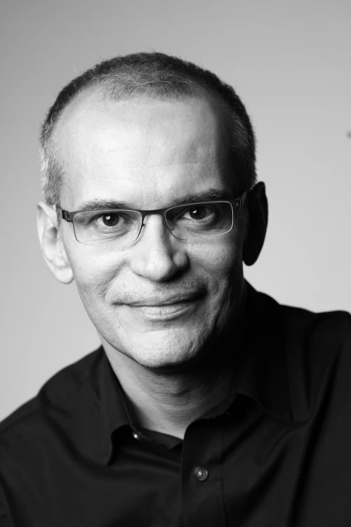Black and white portrait of a middle-aged man with short hair, glasses, and a slight smile, wearing a dark collared shirt, facing the camera against a plain background.